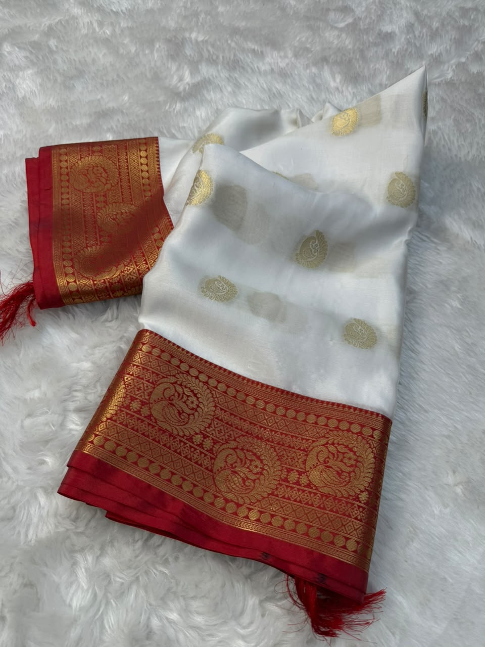 Folded white tapeta saree with gold motifs, red-gold borders, and tassel ends